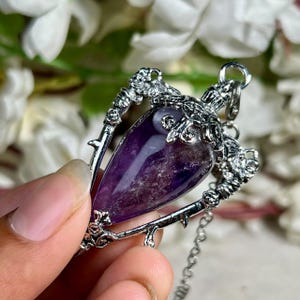 May include: A teardrop-shaped purple amethyst gemstone pendant encased in a silver-toned metal frame. The frame features intricate decorative detailing with small thorns and floral motifs, and includes a small metal loop at the top for a chain.