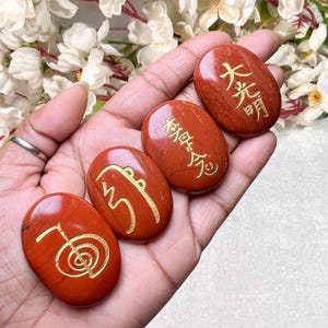 Op de afbeelding: Vier gladde, ovaalvormige rode stenen met gepolijste oppervlakken, elk voorzien van een uniek goudkleurig gegraveerd symbool. De stenen zijn op een rij gerangschikt en tonen kalligrafische tekens en Reiki-gerelateerde emblemen in het materiaal.