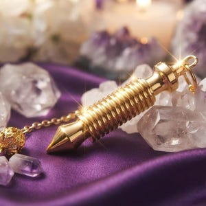 May include: A gold-toned metal pendulum with a ribbed cylindrical body and a pointed tip, attached to a delicate gold chain featuring a small, intricate spherical bead at the end, resting on a smooth purple fabric surface surrounded by clear crystals.