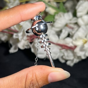 May include: A silver-toned metal pendant shaped like a planet with a ring around its center, featuring a tiered, stepped cylindrical base extending downward and a delicate silver chain attached to the side of the spherical top section.