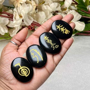 Op de afbeelding: Drie gladde, ovaalvormige zwarte stenen met goudkleurige gegraveerde symbolen. Elke steen heeft een uniek ontwerp, waaronder een spiraalvormig embleem en twee verschillende sets Japanse kanji-tekens, rustend in een handpalm.
