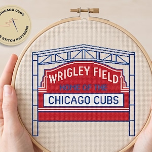 May include: A cross stitch pattern in a wooden embroidery hoop featuring the Wrigley Field marquee. The design uses red, white, and blue thread on cream fabric, with the text "WRIGLEY FIELD HOME OF THE CHICAGO CUBS" clearly stitched in the center.