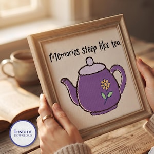 May include: A framed cross-stitch embroidery featuring a purple teapot with a yellow flower design and the text Memories steep like tea in black lettering. The piece is set within a light-colored wooden frame and displayed on a white fabric background.