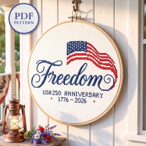 Op de afbeelding: Een ronde borduurring met een kruissteekontwerp op witte stof. Het ontwerp toont een gestileerde Amerikaanse vlag, het woord Freedom in blauw schrift en de tekst USA 250 ANNIVERSARY 1776 - 2026 met kleine rode kruisjes.