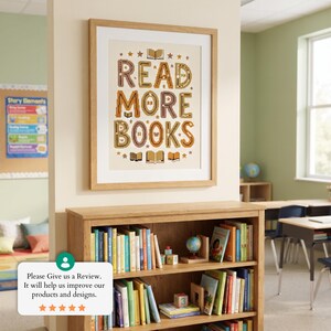 May include: A framed wall art print featuring the text READ MORE BOOKS in a brown, tan, and cream color palette. The design includes small star and open book icons surrounding the lettering, all encased in a light-toned wooden frame.