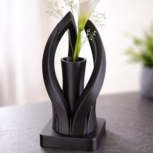 Könnte beinhalten: Eine moderne schwarze Vase mit einem einzigartigen, offenen Design, die eine weiße Calla-Lilie und zarte weiße Füllblumen enthält. Die Vase hat einen quadratischen Sockel und einen zylindrischen Innenraum.