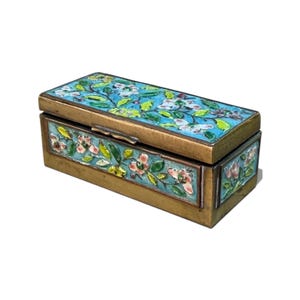May include: A rectangular brass box featuring a hinged lid and sides decorated with colourful enamel floral patterns. The design displays light blue backgrounds with green leaves and pink and white flowers, set against a metallic brass frame.