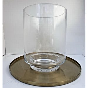 Peut inclure: Un vase cylindrique en verre transparent avec une large ouverture, posé sur un plateau rond de couleur laiton antique. Le vase a une base épaisse et un design simple et élégant. Le plateau mesure environ 30 cm de diamètre.