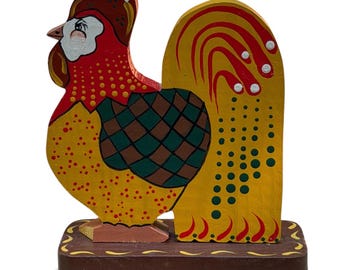 May include: A hand-painted wooden rooster figurine standing on a rectangular base. The rooster features a red head, a yellow body with red and green polka dots, and a green and brown checkered wing. The tail is yellow with red and green accents.