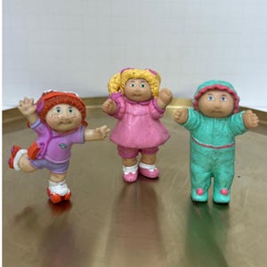 Könnte beinhalten: Drei Vintage-Figuren von Cabbage Patch Kids. Eine trägt einen roten Hut und ein lila Outfit, eine andere ein rosa Kleid und die dritte ein türkisfarbenes Outfit. Alle haben die klassischen Gesichtszüge der Cabbage Patch Kids.