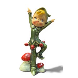 May include: A ceramic figurine of a cheerful elf with blonde hair, wearing a dark green outfit with red accents, standing on two red and white mushrooms. The elf has arms raised in a joyful pose, with a glossy finish and painted facial features.