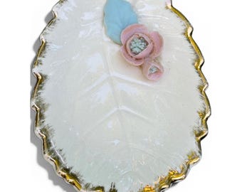 May include: A white ceramic dish shaped like a leaf with a serrated edge trimmed in gold. The surface features a raised, light blue leaf accent and a small, three-dimensional pink flower with a pale green center attached to the top right side.