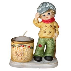 May include: A ceramic figurine of a young boy in a yellow coat, green pants, and a red scarf, holding a cap. Next to him is a candle holder with the text "God Bless Us Everyone". The candle holder is decorated with holly berries.