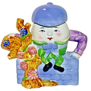 Könnte beinhalten: Keramikfigur von Humpty Dumpty, mit blauer Mütze, weißem eiförmigen Körper und lächelndem Gesicht. Er trägt eine grüne Jacke, einen lila Ärmel und schwarze Schuhe und sitzt auf einer blauen Backsteinmauer mit einem Blumenkorb.