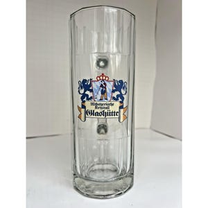 May include: A tall, clear glass beer stein with a handle. The stein features a decorative emblem with a crest, two blue lions, and the text "Altbayerische Kristall Glashütte."