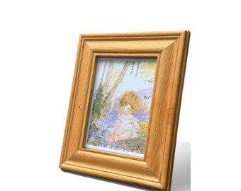 May include: A rectangular wooden picture frame with a light brown, natural wood grain finish. The frame holds a print featuring a watercolor-style illustration of a child with curly blonde hair sitting outdoors, accompanied by a small yellow bear.