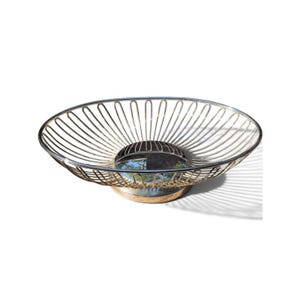 Puede incluir: Un cuenco de alambre metálico en tono plateado con un recipiente ovalado, ancho y poco profundo, construido con finas varillas verticales espaciadas uniformemente que convergen en una base circular sólida, creando una estructura reflectante.
