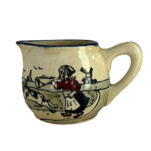 May include: Cream-colored ceramic creamer with a blue rim and handle. The creamer features a hand-painted scene of a person playing a horn, a dog, and a windmill. The design is in shades of red, black, and blue.