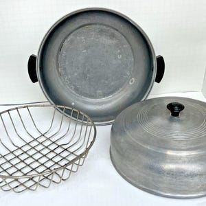 May include: A vintage aluminum roasting pan set with a lid, a wire basket, and a large pan. The lid has a black knob. The pan has black handles. The set is made of aluminum.