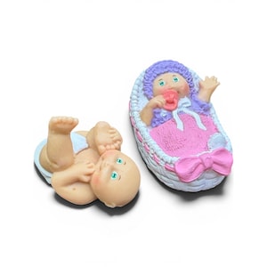 May include: Two small baby figurines. One figurine is lying on its back with legs raised, wearing a white diaper. The other figurine is inside a white oval cradle with a pink blanket, a purple bonnet, and a pink bow, holding a pink pacifier.