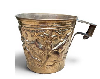 May include: A metallic brass cup featuring an embossed relief design of palm trees and elephants around the exterior surface. The vessel has a wide, open top rim and a single, angular metal handle attached to the side with a visible rivet detail.