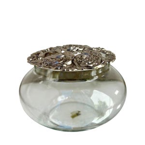 May include: A clear glass jar with a silver-colored lid. The lid is decorated with a floral design of roses. The jar has a round shape and is sitting on a white background.