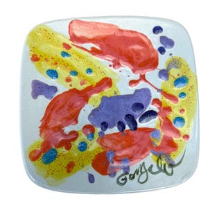 May include: A square glass plate with rounded corners featuring a white speckled base. The surface is decorated with abstract, fluid brushstrokes in vibrant red, yellow, and purple, accented by small blue dots and a dark green signature in the corner.