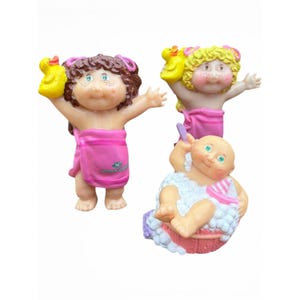 May include: Three small plastic figurines of babies. Two babies have brown and yellow hair, wearing pink towels and holding yellow rubber ducks. The third baby is bald, sitting in a pink tub filled with white bubbles, holding a small pink boat.