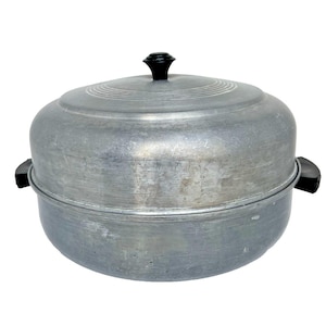 Puede incluir: Una vaporera vintage de aluminio con tapa y asas negras. La olla tiene forma redonda con una superficie ligeramente texturizada. La tapa tiene un pomo negro en la parte superior.