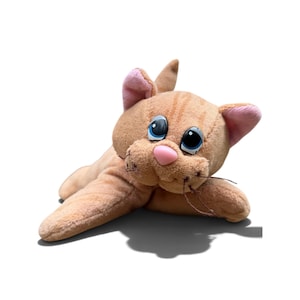 May include: A plush toy cat with light orange fur and darker orange stripes. The cat has large blue plastic eyes, a small pink plastic nose, and pink inner ears. It features thin brown whiskers and is in a lying down position.
