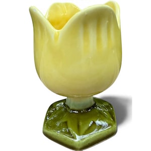 May include: A ceramic egg cup shaped like a yellow tulip flower with a glossy finish. The base of the cup is designed as a dark green leaf structure, providing a stable foundation for the tulip-shaped bowl that holds the egg securely in place.