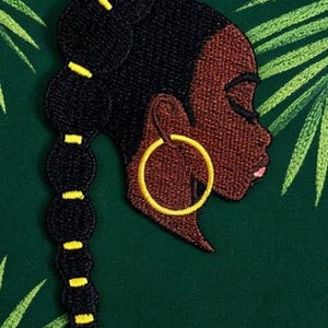 May include: An embroidered patch featuring the profile of a person with dark brown skin, black hair styled in a long bubble ponytail with yellow accents, and a large gold hoop earring. The design is rendered in detailed thread work.