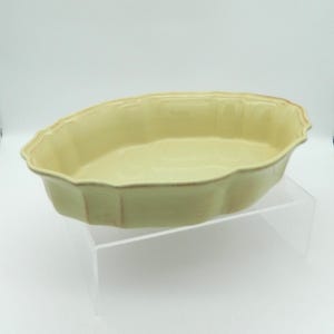May include: A light yellow oval baking dish with a scalloped edge. The dish has a slightly distressed look with brown accents around the rim. The dish is made of ceramic and is on a clear acrylic stand.