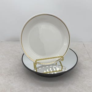 May include: White ceramic plate with a gold rim, resting on a black mirrored surface. The plate is held by a gold-colored wire stand. The design is simple and elegant, suitable for serving food or as a decorative item.