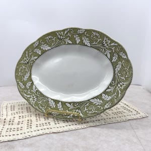 May include: Oval serving platter with a white center and a green and white floral design around the rim. The platter has a scalloped edge and is displayed on a gold-colored stand.