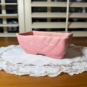 May include: A rectangular pink ceramic planter featuring a subtle, raised white splatter pattern across its exterior. The vessel has a slightly flared rim, a smooth finish, and a molded, rippled texture along the sides, providing a distinct look.