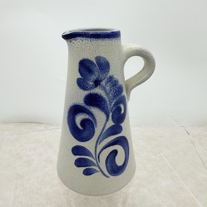 May include: A ceramic pitcher with a cream-colored base and a blue floral design. The pitcher has a handle and a spout. The rim and the floral design are in a deep blue color. The pitcher is likely used for serving beverages.