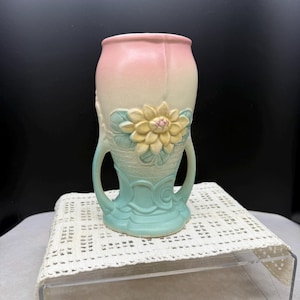 May include: A ceramic vase with a two-handled design. The vase features a gradient of colors, transitioning from pink to cream and ending in a light blue base. A yellow flower with green leaves is embossed on the front.