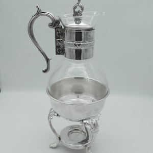 May include: A silver-plated coffee or tea server with a clear glass carafe. The server has a decorative handle and a lid with a swirl design. The carafe sits on a silver stand with ornate legs and a small warming area.