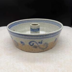 May include: A round ceramic baking dish with a central chimney, featuring a speckled off-white glaze, a tan unglazed base, and blue hand-painted swirl patterns around the exterior and a blue band around the interior rim and the central tube.