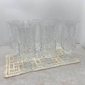 May include: A set of eight clear, stemmed drinking glasses with a detailed, etched leaf pattern. The glasses are arranged on a cream-colored, crocheted placemat. The glasses have a wide bowl and a pedestal base.