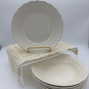 May include: A set of white ceramic plates with a scalloped edge and embossed floral design. One plate is displayed on a gold-colored plate stand, while the others are stacked below. The plates have a cream-colored appearance.