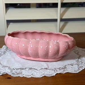 May include: An oval-shaped ceramic planter with a scalloped rim and a ribbed exterior. The piece features a glossy pink glaze finish accented with a white splatter pattern throughout the surface, creating a textured appearance.