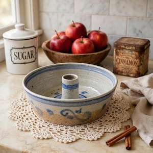 May include: A round ceramic baking pan with a central tube, featuring a speckled beige finish and blue painted scroll patterns. The pan sits on a cream-colored crocheted doily, surrounded by a sugar canister, a bowl of red apples, and cinnamon sticks.