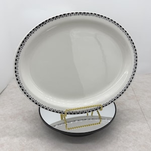 May include: Oval white serving platter with a black decorative border. The platter is displayed on a gold-colored metal stand with a black base. The border design consists of small, repeating black shapes.