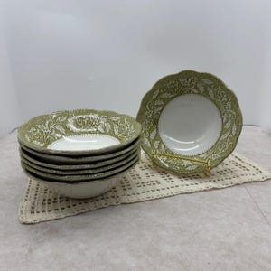 May include: A set of six white and green ceramic bowls with a floral design. The bowls have a scalloped edge and are stacked on a cream-colored crocheted placemat. One bowl is displayed on a gold-colored stand.