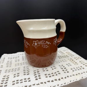 May include: A ceramic pitcher featuring a two-tone design with a cream-colored upper section and a dark brown lower section. The brown portion is decorated with a white etched floral pattern. It includes a curved handle and a pouring spout.