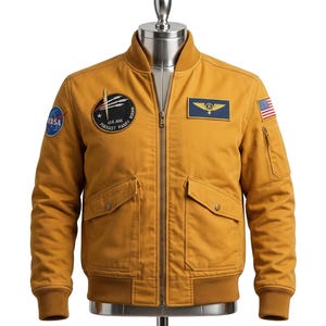 May include: Mustard-coloured bomber jacket with a ribbed collar and cuffs. Features a NASA patch, a US flag patch, and a blue and gold patch. Includes two front pockets and a zip closure. The jacket is displayed on a mannequin.