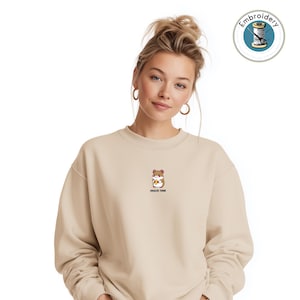 Pode incluir: Camisola bege com um pequeno desenho bordado de um hamster segurando uma banana. O texto "SNACK TIME" está impresso abaixo do desenho. A camisola tem gola redonda e mangas compridas.