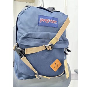 May include: A blue Jansport backpack with tan straps and a brown leather patch. The backpack has a front pocket and a rectangular patch with the brand name. The straps have black plastic buckles.